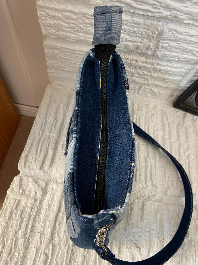May include: A shoulder bag constructed from various shades of blue denim fabric patches. The bag features a top zipper closure with a black zipper track, a single denim strap attached with silver-toned metal hardware, and a visible interior lining.