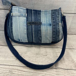 May include: A shoulder bag constructed from vertical strips of denim in varying shades of blue, ranging from light wash to dark indigo. The bag features a single dark denim strap attached with silver-toned metal hardware and a top zipper closure.