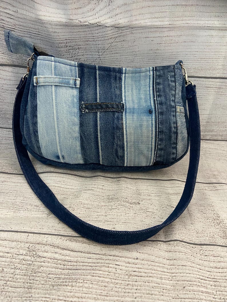 May include: A shoulder bag constructed from vertical strips of denim in varying shades of blue, ranging from light wash to dark indigo. The bag features a single dark denim strap attached with silver-toned metal hardware and a top zipper closure.