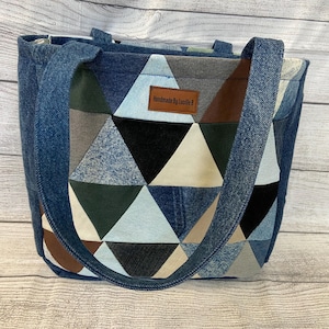 May include: A denim tote bag with a geometric patchwork design. The patchwork features triangles in shades of blue, black, gray, and brown. The bag has two handles and a label that reads "Handmade by Lucille B."