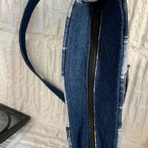May include: A shoulder bag constructed from various shades of blue denim fabric. The bag features a central black zipper closure along the top edge and a matching dark blue denim shoulder strap. The interior lining displays a floral pattern.