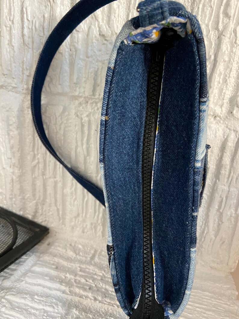 May include: A shoulder bag constructed from various shades of blue denim fabric. The bag features a central black zipper closure along the top edge and a matching dark blue denim shoulder strap. The interior lining displays a floral pattern.