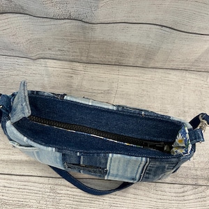 May include: A small handbag constructed from various shades of blue denim fabric in a patchwork design. The bag features a black zipper closure along the top opening and a blue denim strap attached with silver-toned metal hardware on the sides.