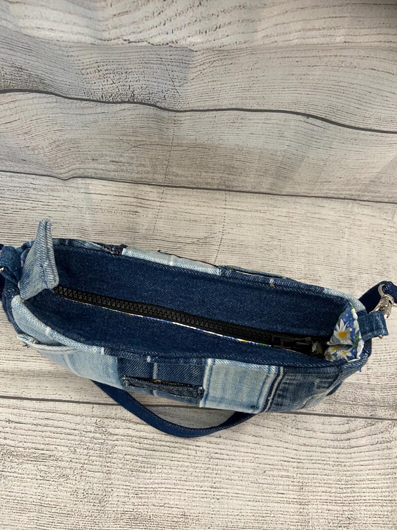 May include: A small handbag constructed from various shades of blue denim fabric in a patchwork design. The bag features a black zipper closure along the top opening and a blue denim strap attached with silver-toned metal hardware on the sides.