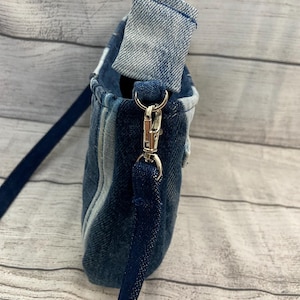 May include: A crossbody bag constructed from various shades of blue denim fabric. The bag features a folded top edge, a silver-toned metal swivel clasp, and a dark blue fabric strap attached to the side of the bag with a reinforced denim loop.