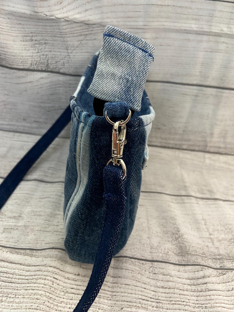 May include: A crossbody bag constructed from various shades of blue denim fabric. The bag features a folded top edge, a silver-toned metal swivel clasp, and a dark blue fabric strap attached to the side of the bag with a reinforced denim loop.