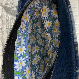 May include: A denim handbag featuring a dark blue exterior and a contrasting interior lining decorated with a repeating pattern of white daisies with yellow centers on a light blue background, secured by a black zipper closure.