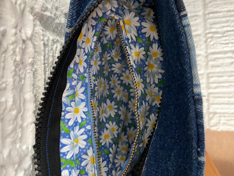 May include: A denim handbag featuring a dark blue exterior and a contrasting interior lining decorated with a repeating pattern of white daisies with yellow centers on a light blue background, secured by a black zipper closure.