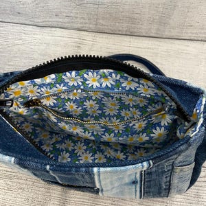 May include: A denim crossbody bag featuring a patchwork exterior made of various shades of blue fabric. The interior is lined with a blue fabric patterned with white daisies and yellow centers, including a zippered pocket with a gold-toned metal zipper.