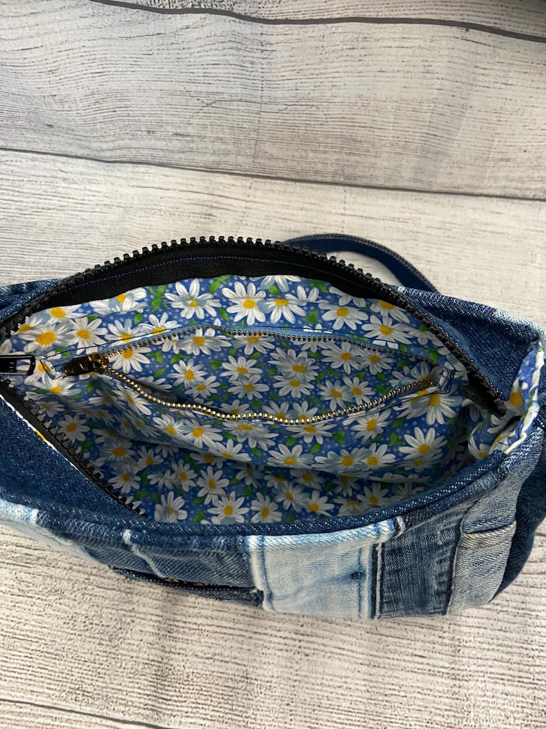May include: A denim crossbody bag featuring a patchwork exterior made of various shades of blue fabric. The interior is lined with a blue fabric patterned with white daisies and yellow centers, including a zippered pocket with a gold-toned metal zipper.
