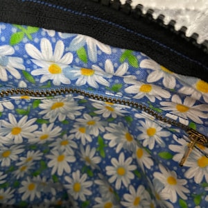 May include: The interior of a bag featuring a blue fabric lining with a repeating pattern of white daisies with yellow centers and small green leaves. A metallic zipper runs horizontally across the middle, and a black zipper is visible at the top.