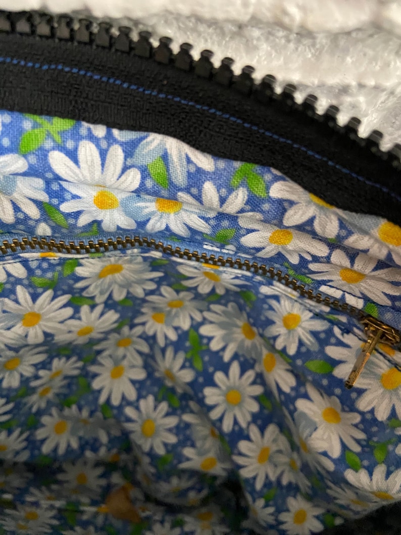 May include: The interior of a bag featuring a blue fabric lining with a repeating pattern of white daisies with yellow centers and small green leaves. A metallic zipper runs horizontally across the middle, and a black zipper is visible at the top.