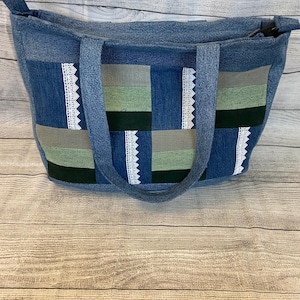 May include: A blue denim tote bag featuring a patchwork design of horizontal stripes in shades of gray, sage green, and dark forest green. The bag is accented with vertical strips of white lace trim and includes two matching denim shoulder straps.