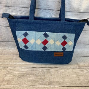 May include: A dark blue denim tote bag featuring a horizontal band with three quilted square patterns in light blue, dark blue, white, and red. The bag has two top handles, a zipper closure, and a brown rectangular label reading Handmade By Lucille P.