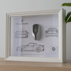 May include: A white frame with a white mount displaying a black and white technical drawing of a Maserati car. The drawing is accompanied by a silver key fob with the Maserati trident logo. The frame also includes the Maserati logo and the text "Maserati".