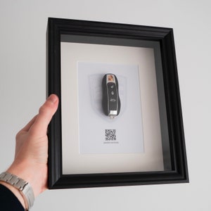 Peut inclure: Cadre noir avec passe-partout blanc. À l'intérieur, une clé de voiture noire et argentée est exposée sur un fond blanc avec un emblème Porsche. Les mots "LIMITED EDITION" sont imprimés sous un code QR.