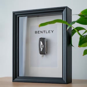 May include: A black framed shadow box with a white mat and a silver car key with the Bentley logo. The key is hanging from a silver chain. The text "BENTLEY" is printed on the mat.