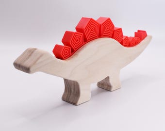 May include: A wooden dinosaur toy with a red, 3D printed, spiky back. The dinosaur is standing on a white surface.
