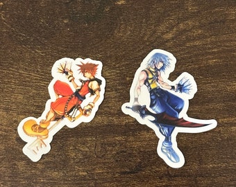 Könnte beinhalten: Zwei bunte Aufkleber mit Figuren aus einem beliebten Videospiel. Der Aufkleber links zeigt eine Figur mit orangefarbenem Haar, einem rot-gelben Outfit und einer Keyblade. Der Aufkleber rechts zeigt eine Figur mit blauen Haaren, einem blau-gelben Outfit und einem Schwert.