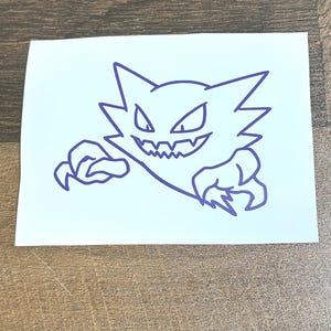 May include: A purple outline of a cartoon ghost with sharp teeth and claws. The ghost has a jagged head and is on a white rectangular sticker. The sticker is on a wooden surface.