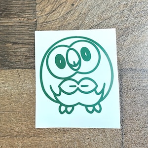 May include: A green owl sticker on a white background. The owl has large eyes, a beak, and a stylized feather design. The sticker is rectangular and sits on a wooden surface.