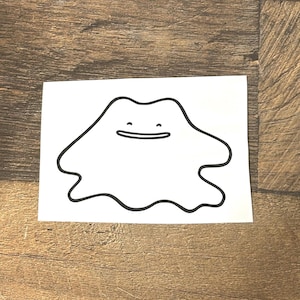 May include: A white sticker featuring a black outline of a Ditto character. The character has a blob-like shape with a smiling mouth. The sticker is on a wooden surface.