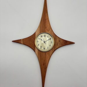 May include: A mid-century modern starburst clock. The clock features a wooden starburst design with a round clock face in the center. The clock face has black numbers and hands with a gold-colored bezel. The wood has a natural finish.