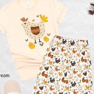 May include: A soft cream colored t-shirt with a cartoon illustration of a hen and chicks. The t-shirt is paired with white pajama pants with a repeating pattern of hens and chicks.