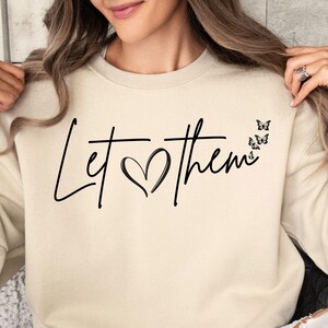 May include: A beige sweatshirt with the text "Let &hearts; them" in black script with a heart and four small black butterflies. The text is centered on the sweatshirt. The sweatshirt is being worn by a person sitting down. The color of the sweatshirt is "Sand".