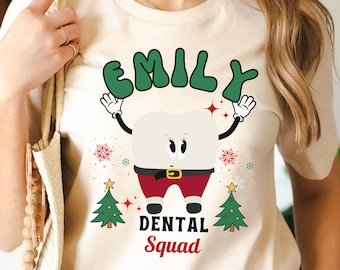 May include: A cream-colored t-shirt featuring a cartoon tooth character dressed in a Santa Claus outfit. The text "EMILY" is arched above the tooth, with "DENTAL Squad" below. Christmas trees and snowflakes decorate the design.