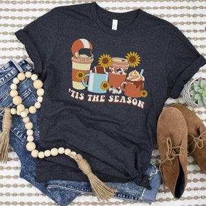 May include: A dark blue t-shirt with a graphic of a football, coffee cups, and sunflowers. The text "'Tis the season" is printed on the shirt.