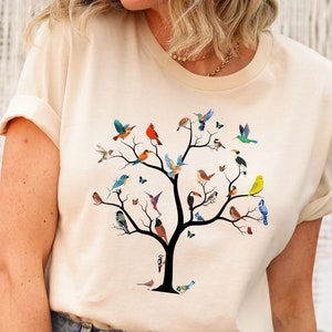 May include: A beige t-shirt with a colorful tree design featuring a variety of birds. The tree has a brown trunk and branches with green leaves. The birds are in various colors, including red, blue, green, yellow, and orange.
