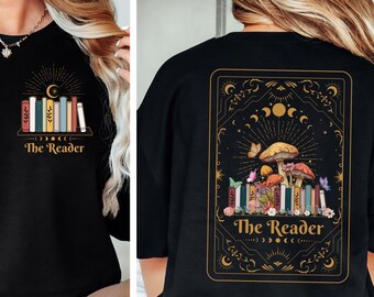 Pode incluir: Camiseta preta com um design de carta de tarô dourado que apresenta uma pilha de livros, cogumelos e o texto "The Reader".