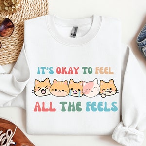Puede incluir: Sudadera blanca con el texto "It's okay to feel all the feels" en colores arcoíris. Cinco gatos de dibujos animados con diferentes expresiones faciales están alineados debajo del texto.