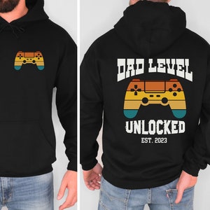 May include: Black hoodie with a retro-style video game controller graphic on the front and the text "DAD LEVEL UNLOCKED EST. 2023" on the back.