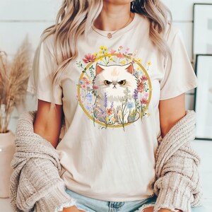 May include: A white t-shirt with a graphic of a white fluffy cat with a grumpy expression surrounded by colorful flowers in a gold circle.