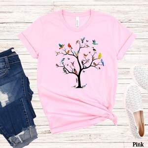 May include: A pink t-shirt with a colorful bird tree design. The tree has a black trunk and branches with many different colored birds perched on the branches.