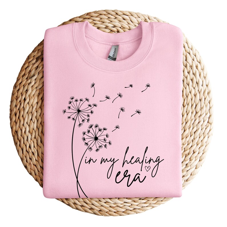 K&ouml;nnte beinhalten: Rosafarbener Pullover mit einem schwarzen L&ouml;wenzahnmuster und dem Text "in my healing era" mit einem kleinen Herzen.