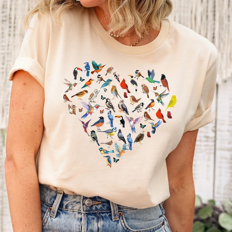 May include: A cream-coloured t-shirt with a heart-shaped design featuring a variety of colourful birds. The birds are arranged in a heart shape and are all different sizes and species.
