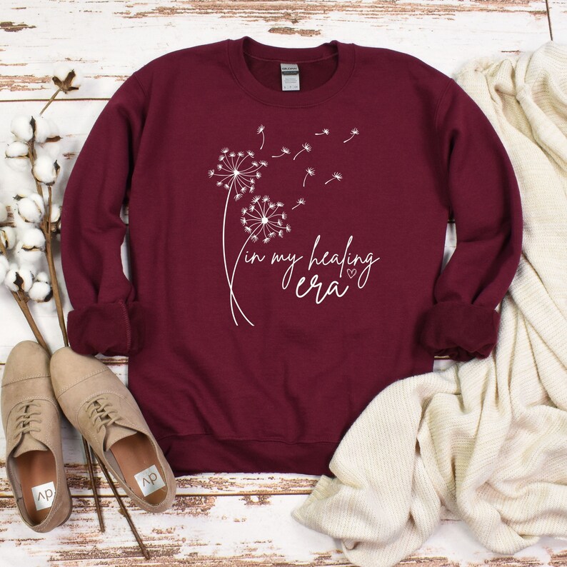K&ouml;nnte beinhalten: Burgunderfarbener Pullover mit einem wei&szlig;en L&ouml;wenzahnmuster und dem Text "in my healing era" mit einem kleinen Herzen.