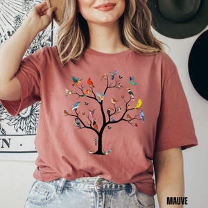 May include: A mauve t-shirt with a graphic of a tree with many colorful birds on the branches. The text "MAUVE" is in the lower right corner of the image.
