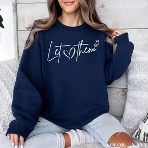 May include: Navy blue sweatshirt with the text "Let them" in white with a heart symbol. The sweatshirt has a relaxed fit and long sleeves.
