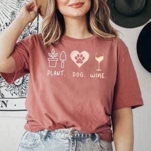 Pu&ograve; includere: Una donna indossa una maglietta color granata con un design grafico di una pianta, un'impronta di zampa di cane all'interno di un cuore e un bicchiere di vino. Il testo sotto la grafica recita "PLANT. DOG. WINE."