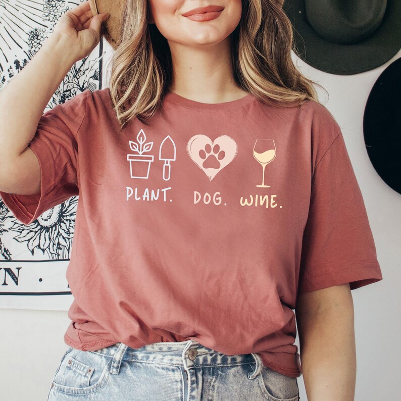 Pu&ograve; includere: Una donna indossa una maglietta color granata con un design grafico di una pianta, un'impronta di zampa di cane all'interno di un cuore e un bicchiere di vino. Il testo sotto la grafica recita "PLANT. DOG. WINE."