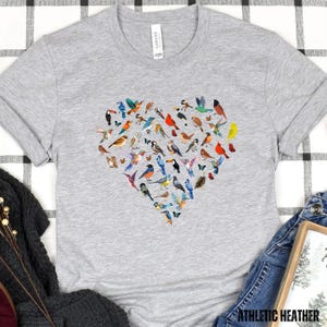 May include: A grey t-shirt featuring a heart-shaped design made up of colourful birds. The birds are arranged in a heart shape and are all different colours and sizes.