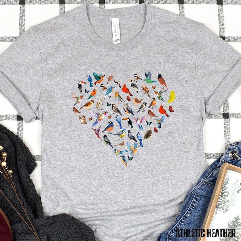 May include: A grey t-shirt featuring a heart-shaped design made up of colourful birds. The birds are arranged in a heart shape and are all different colours and sizes.