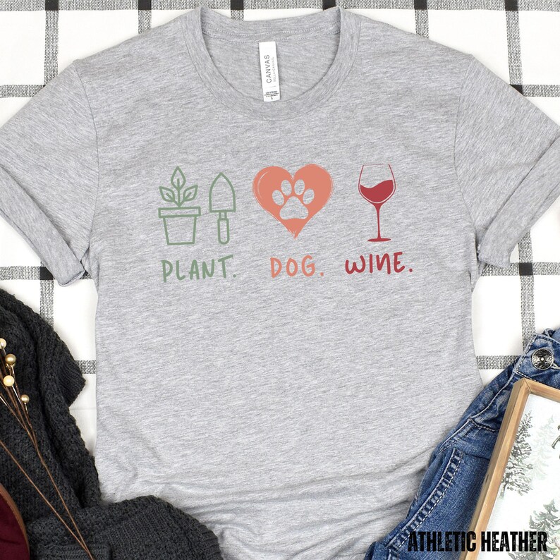 Pu&ograve; includere: Maglietta grigia con un design grafico di una pianta, un'impronta di zampa all'interno di un cuore e un bicchiere di vino. Il testo "PLANT. DOG. WINE." &egrave; stampato sotto il grafico.