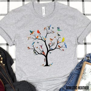 May include: A gray t-shirt with a colorful tree design featuring a variety of birds. The tree has a dark brown trunk and branches with green leaves. The birds are in various colors, including blue, red, yellow, green, and orange.