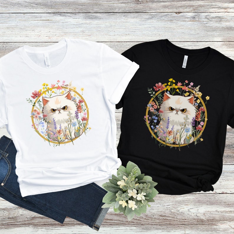 May include: Two t-shirts with a white Persian cat with angry eyes surrounded by colorful flowers in a gold circle. The cat is on a white t-shirt and a black t-shirt.