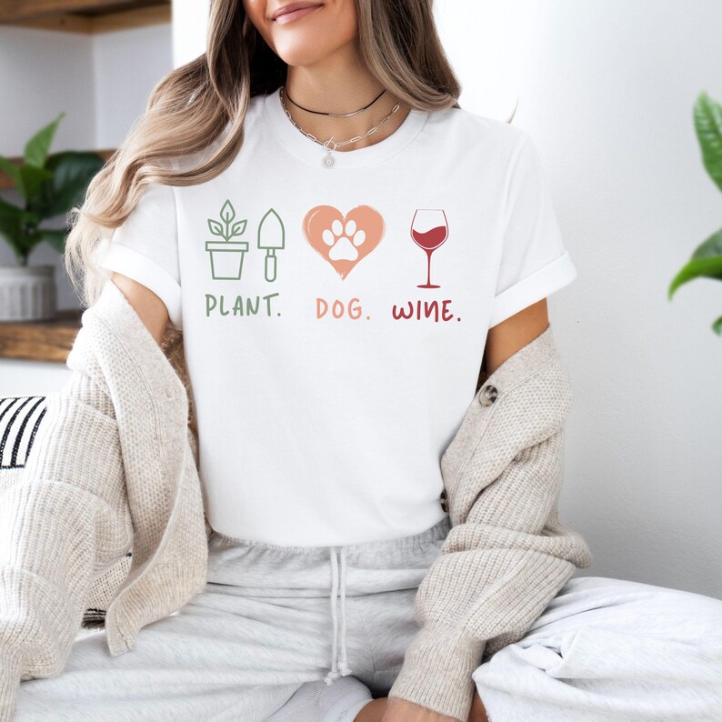 Pu&ograve; includere: Maglietta bianca con un design grafico che presenta una pianta, un'impronta di zampa di cane all'interno di un cuore e un bicchiere di vino. Il testo sotto la grafica dice "Plant. Dog. Wine."